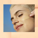 Garnier BB Cream All In One Perfector Classic Light SPF 15  - Close-up of a person with short platinum blonde hair applying a swipe of beige liquid foundation or concealer to their cheek against a blue sky background, framed by a peach border.