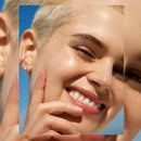A close-up lifestyle shot of a smiling woman with a buzz cut outdoors under a blue sky, wearing a small silver hoop earring and a stack of silver earrings. Garnier BB Cream All In One Perfector Classic Light SPF 15 