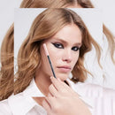 A close-up of a woman with blonde hair holding a pink and black makeup pencil near her face, wearing a white collared shirt against a light gray background.