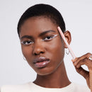L'Oreal Paris Paradise Le Khol Eyeliner 120 Immaculate Snow - A close-up of a model with short dark hair wearing dramatic white winged eyeliner and holding a light pink lip liner pencil against a clean light gray background.