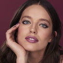 Close-up portrait of a woman with long dark brown hair, blue eyes, and mauve-purple lipstick, resting her hand near her face against a solid dark burgundy background.