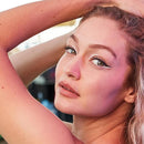 A close-up portrait of a woman with white winged eyeliner, looking towards the camera with her arms raised, set against a softly blurred background. Maybelline Tattoo Gel Pencil Eyeliner